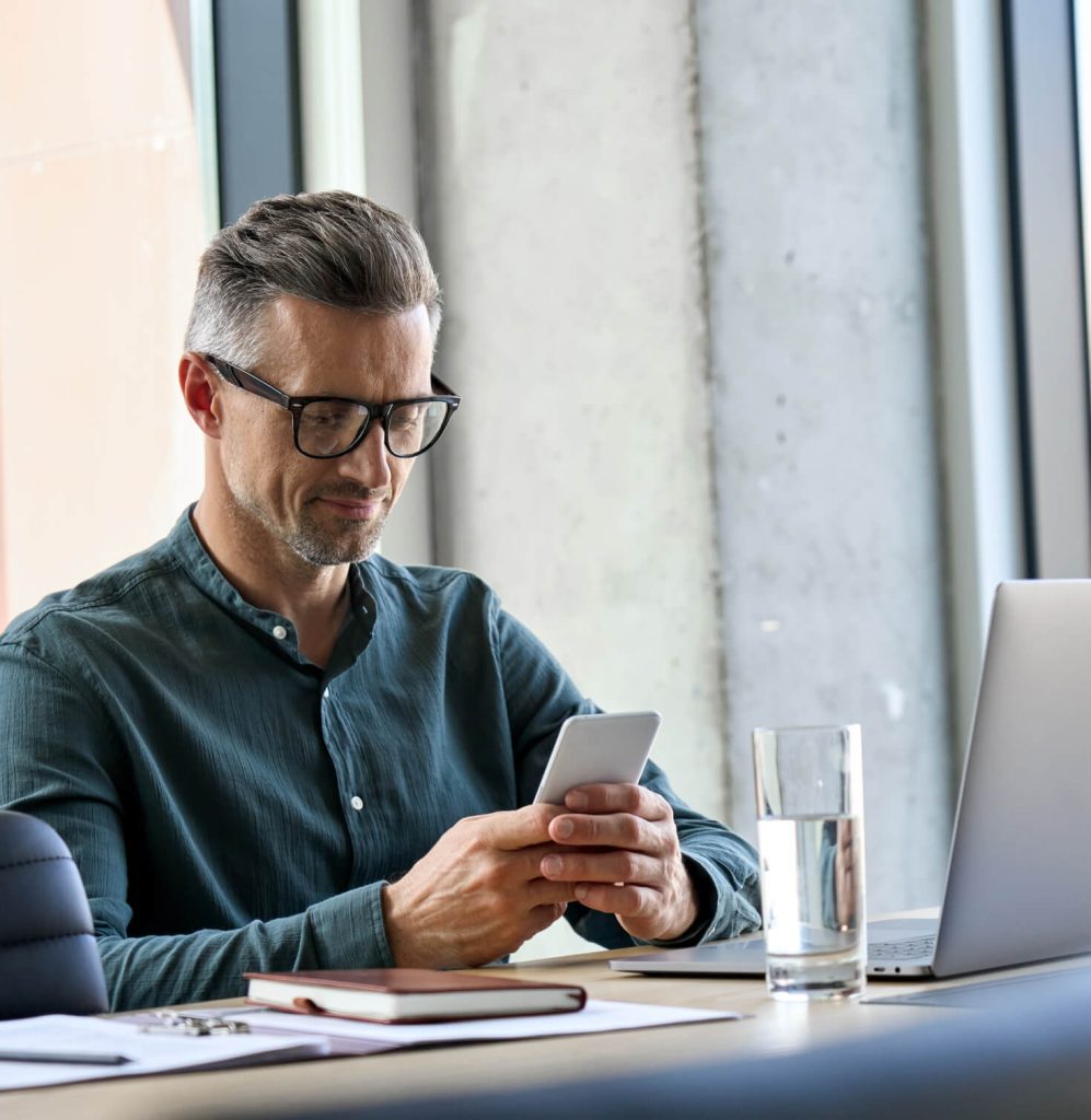 man with glasses typing on a cell phone next to his computer